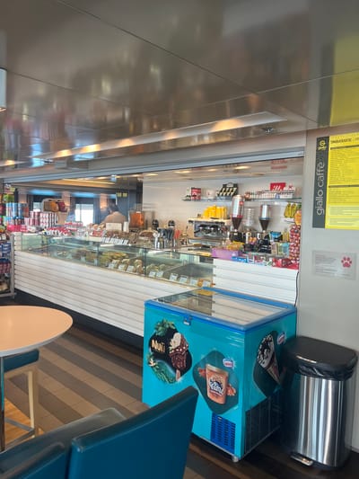 The image depicts the interior of a cafe or convenience store, likely onboard a ferry or in a transportation hub, given the presence of packaged goods, a refrigerated display case, and a coffee station. The overall aesthetic is clean and modern, with metallic and white surfaces dominating the upper half of the image, suggesting a functional and possibly high-traffic environment.

In the foreground, a round, light-colored table is partially visible, with a glimpse of blue seating. To the right, a bright blue, chest-style freezer with imagery of ice cream cones and bars is prominently displayed. Next to the freezer, a stainless steel trash can with a black lid and pedal is positioned. The floor is tiled in a geometric pattern of dark grey and brown stripes.

The mid-ground is dominated by a long, refrigerated display case filled with various food items, including sandwiches, pastries, and possibly pre-packaged snacks. Behind the display case, a coffee machine with grinders and dispensers is visible, along with shelves stocked with coffee-related products and other packaged goods. A man in a light-colored shirt is standing behind the counter, seemingly attending to customers or preparing items.

The background features a reflective ceiling and more shelving units stocked with a variety of merchandise, including snacks, drinks, and possibly toiletries or travel essentials. A yellow sign with Greek text and the "giallo caffè" logo is mounted on the right wall. The lighting appears to be a mix of overhead fluorescent lights and accent lighting, creating a well-lit but somewhat sterile atmosphere. The presence of the "giallo caffè" branding and the menu in Greek strongly suggests a location in Greece. Considering the context of transportation hubs and the specific signage, the Municipal Unit of Kastro - Kyllini, Greece, which includes the port of Kyllini, a major ferry terminal, is a highly probable location for this establishment.