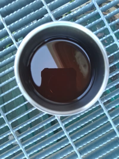 A close-up, overhead view captures a metallic mug filled with a dark liquid, likely coffee or tea, resting on a grey metal grate. The grate appears to be part of an outdoor surface, possibly a walkway or seating area, with hints of green foliage visible through the gaps in the metal. The liquid in the mug reflects a dark, rectangular object, possibly a smartphone, suggesting the presence of a person taking the photograph. The setting appears to be outdoors, with natural light illuminating the scene. There is no information in the image that places this scene in Nicorești, Romania.