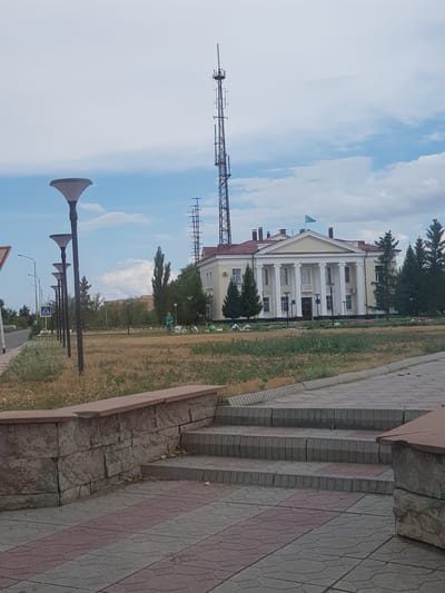 In Kurchatov, Kazakhstan, a large, multi-story building with a classical facade featuring a prominent portico with white columns stands under a cloudy sky. A light blue flag flies from the roof of the building. Adjacent to the building, a tall, lattice-like telecommunications tower dominates the skyline. In front of the building, a manicured lawn is interspersed with trees, and several people are visible, some appearing to be engaged in gardening or maintenance. A paved walkway with decorative red and gray tiles leads up to a set of stone steps. Several streetlights with distinctive conical lamps line the walkway. The scene suggests a public space or government building in a suburban or semi-urban environment.