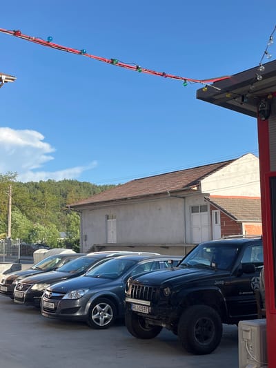 The image shows a partially obscured view of a street in Satovcha, Bulgaria, featuring a row of parked cars. The vehicles, including a Jeep Cherokee with license plate PK 1435 BT, are parked along a concrete surface resembling a parking lot or roadside. In the background, there's a building with a red-tiled roof and a lighter-colored exterior, nestled amidst trees. A string of lights hangs on the left side of the image against a blue sky with scattered clouds.