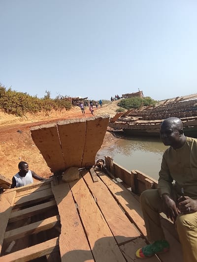 The image depicts a daytime scene in Ibi, Nigeria, at what appears to be a riverbank or construction site for wooden boats. The sky is clear and light blue. The terrain is composed of reddish-brown dirt, with sparse green vegetation visible on elevated banks.
In the foreground, an adult male with dark skin and short dark hair is seated on the raw wooden planks of a boat deck, positioned to the right. He wears a long-sleeved olive-green tunic and trousers. He holds a dark, thin object in his left hand and wears a ring on the ring finger of his right hand. A pair of green and yellow sandals rests on the deck directly in front of him. He is looking towards the right, out of frame, with a slight smile.
To the left of this individual, on the same boat, a tall, upright wooden structure, possibly the bow section, rises from the deck. A light-colored rock is placed on the deck near its base. Partially obscured behind this structure, another adult male with dark skin and short dark hair is visible, wearing a white sleeveless top. He appears to be interacting with the boat's interior or structure.
In the mid-ground, a body of water, likely a river, is visible to the right of the foreground boat. Several other wooden boat structures, some appearing to be partially submerged or under construction/repair with visible wooden framing, are present in the water and along the bank.
In the background, a dirt path or road ascends an embankment. Multiple individuals are visible on this path and on the higher ground, appearing to be walking or gathered. Several rudimentary structures are also discernible on the distant bank.