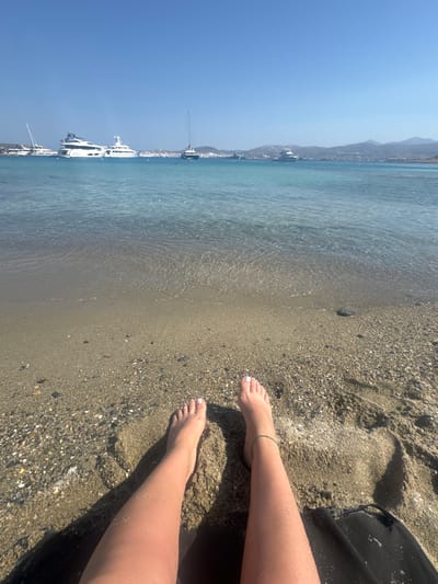 A person's legs and feet rest on a sandy beach next to the clear turquoise water of the Aegean Sea. The image is taken from a first-person perspective, looking down at the scene. The person appears to be relaxing, with one leg extended and the other bent. A black object, possibly a beach towel or mat, is partially visible to the right. In the background, several white yachts are anchored in the bay, with a coastal town and hills visible on the horizon. The sky is a clear blue, suggesting a sunny day. The water is calm with gentle ripples. The sand is a mix of fine grains and small pebbles. The overall impression is one of a peaceful and idyllic beach vacation. The time of day appears to be midday, given the bright sunlight and clear shadows. The location cues of the yachts and the coastal landscape are consistent with a Mediterranean setting, specifically Naousa, Greece. There is no visible text in the image.