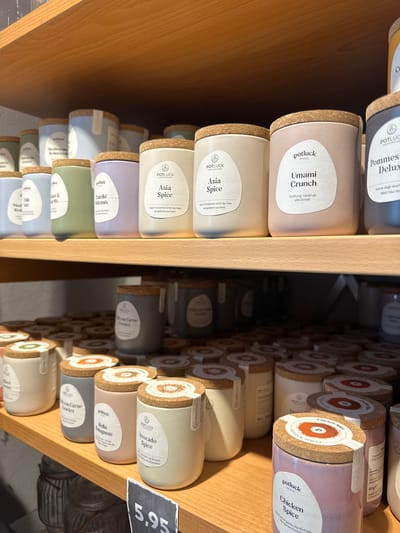 This image shows a close-up view of shelves stocked with small spice jars. The jars are mostly cylindrical with cork stoppers, and each has a label indicating the spice blend inside. The labels are predominantly white with text in various colors, including black, green, and red. Some of the visible spice names are "Pommes Deluxe," "Umami Crunch," "Asia Spice," "Tzatziki Würzmix," "Mediterranean Kräuterea," "Chili Med Salz Kra," "Chicken Spice," "Bella Bolognese," and "Avocado Spice." The background is a wooden shelf and a dark, out-of-focus area suggesting a store or pantry setting. A price tag of "5,95" is visible on the right side of the image, further indicating a retail environment. The lighting appears to be artificial, casting shadows and highlighting the textures of the jars and cork. The scene is static, focusing solely on the organization and display of the spice products. There are no people or animals visible. The overall impression is one of an organized retail display, likely in a grocery store or specialty food shop in Herford, Germany.