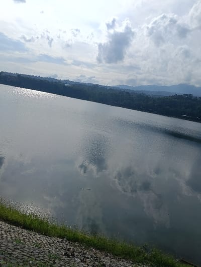 This image displays the Gembong Reservoir in Gembong, Indonesia, viewed from a slightly elevated position under a partly cloudy sky. The foreground, positioned in the lower left, features a sloped bank constructed from dark grey interlocking pavers or cobblestones, transitioning into a narrow strip of green grass at the waterline. The main subject is the expansive water surface of the reservoir, which shows ripples and prominently reflects the overhead clouds. An elongated, bright reflection of sunlight is visible on the water's surface towards the upper left. Several small, dark, elongated objects, possibly natural debris, float on the water near the foreground bank. Across the water, a dense band of dark green foliage indicates a forested shoreline, extending into a series of undulating green hills and distant mountains that form the horizon line. The sky above the landscape contains scattered white and grey cumulus clouds against areas of brighter sky. No individuals, vehicles, or active human structures are identifiable within the scene.