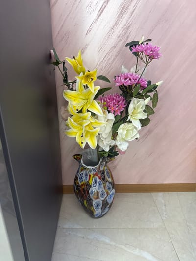 A vase of mixed flowers is arranged on a dark gray shelf in front of a wall. The flowers are a mix of yellow lilies, white roses, and purple allium. The vase is colorful, with an array of blues, yellows, and reds. The wall behind the flowers is pale pink with a brushed texture. To the right of the vase is a corner where the wall changes to a beige marble pattern, separated by a light brown vertical strip. The shelf extends into the foreground, occupying the lower portion of the frame. This scene is likely a staged interior shot, the location in the city of Krichim, Bulgaria is not verifiable from the image.