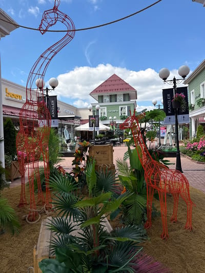 The image depicts an outdoor pedestrian shopping area. Two large, red wireframe giraffe sculptures are prominently displayed among lush green tropical plants and a sandy ground cover. In the background, a row of multi-story buildings with light green facades and red tiled roofs line the walkway. Signs for businesses such as "BOOTW", "Л'Этуаль", "KANZLER", and "COCCINELLE" are visible on the storefronts. Streetlights with white globe lamps are placed along the path, some adorned with black banners. A few tables and chairs are set up outside a restaurant. The sky is blue with scattered white clouds.