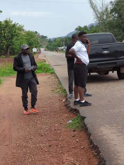 The image shows a scene on the side of a road in Gidan Waya, Nigeria. A black Ford truck is parked on the side of a paved road. Two men stand by the truck; one is talking on a cell phone. Another man is standing on a gravel path near the paved road, holding a cell phone in his hands. The area includes vegetation and an unpaved, reddish-brown gravel path. Other people are seen in the distance.