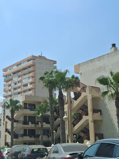 The image depicts a street scene in San Javier, Spain, under a clear blue sky. In the foreground, several cars are parked or driving, including a dark gray hatchback, a silver sedan, and a white car. The midground is dominated by a multi-story apartment building with numerous balconies, featuring a light peach-colored facade with white railings. Adjacent to this building is another structure with a textured off-white exterior and an external staircase with wooden railings. Several palm trees with lush green fronds are scattered throughout the scene, adding to the Mediterranean atmosphere. The buildings appear to be residential or hotels, typical of a coastal town. The overall impression is one of a bright, sunny day in a European city.