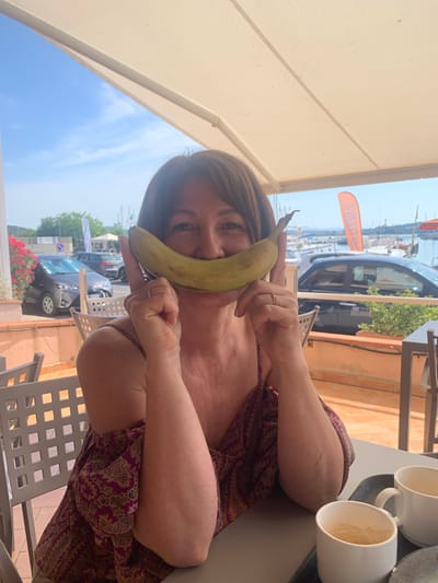 A woman, holding a banana to her face to mimic a smile, sits at a table outdoors. She is wearing a patterned dress. Behind her, a harbor with boats is visible. The scene appears to be in a restaurant or cafe in Orbetello, Italy, given the context provided. The weather is sunny, with a clear blue sky.