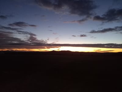 The image displays a wide landscape view captured during either dawn or dusk, located in Santa Juana, Chile. The upper sky is a deep indigo blue, gradually lightening towards the horizon. Scattered dark grey and purplish clouds are present across the upper and middle portions of the sky, with a thin crescent moon visible in the upper left quadrant. Along the horizon, a distinct, vivid band of orange, yellow, and golden light dominates, suggesting the sun's position just below the visible frame. Silhouettes of distant, undulating hills or a dense tree line are discernible against this illuminated horizon. The lower approximately two-thirds of the image comprises a uniformly dark, undifferentiated foreground, consistent with an open field or unlit terrain. There are no individuals, specific objects, or discernible actions present in the foreground or midground.