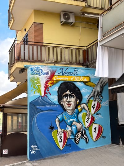 An outdoor mural depicting Argentine football legend Diego Maradona is painted on a light blue wall, which is part of a yellow building. Maradona is shown from the chest up, wearing a light blue Napoli football club jersey with "BUITONI" and his iconic number "10" visible. He appears to be looking directly forward with a determined expression.
Above Maradona, a hand with darker skin tone reaches down, seemingly offering or holding a shield-like object marked with the number "4" and the year "2025". Maradona himself holds a similar shield marked "3" and "2023". Further to the right, two more shield-like objects are depicted: one with "2" and "1990", and another with "1" and "1987". Two black soccer cleats are positioned between the "2" and "1" shields.
The mural features text that reads "Napoli Campione d'Italia" (Napoli Champion of Italy) in elegant script, highlighted in yellow. Below this, there are flame-like motifs in red, yellow, and green, representing the Italian flag. Signatures "Il Dottore Carmine Bianco" and "Claudio Petito - ART" are also present on the mural.
The background shows the exterior of a building with peeling yellow paint and brown brick sections. Several metal balconies with glass panels are visible, along with an outdoor Samsung air conditioning unit attached to the wall. Stairs with similar glass panels ascend along the building's side. The sky, partially visible, is clear blue with white clouds, suggesting daytime. Below the mural, a glimpse of a building entrance with a partially visible "NO SPRAY NO GRAFFITI" sign can be seen on the pavement. The overall mural celebrates Napoli's Serie A titles, including a hopeful prediction for 2025.