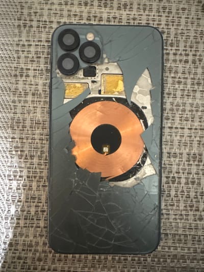The image shows the back of a broken smartphone, likely an iPhone, with its rear glass shattered and peeled away, revealing internal components. The camera module with three lenses is visible at the top. A prominent copper-colored wireless charging coil is exposed in the center. The phone rests on a textured woven surface, possibly a placemat or tablecloth, with a basketweave pattern. The environment appears to be indoors, and there is no indication of the specific location being in Демихово, Russia; this detail is not supported by the visual evidence. The scene depicts a damaged electronic device.