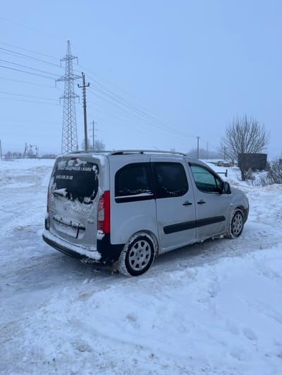A light-colored van, possibly silver, is parked on a snow-covered roadside in Voronino, Russia, under an overcast sky. The vehicle is angled with its rear left side facing the viewer, partially covered in snow and slush. Cyrillic text on the rear window reads "ПРОКАТ КВАДРИКОВ Г. КЛИН" (Quad Bike Rental Klin City) followed by a phone number "8 903 008 30 88". A Russian license plate, "A 494 YX 750", is affixed to the rear. In the background, a large metal lattice electrical transmission tower and several utility poles support multiple power lines extending across the winter landscape. The ground is uniformly covered in snow, with visible tire tracks on the unpaved surface. To the right, a leafless tree and a dark, low structure are partially obscured by snow. No individuals are present in the scene.