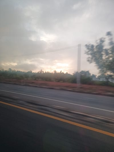 The photograph appears to be taken from a moving vehicle on a road in Surat Thani City Municipality, Thailand. The road is paved with a yellow line on the side. There's vegetation on the roadside with trees and foliage. The sky is overcast, with some sunlight breaking through the clouds. There are power lines running above the trees. The image has motion blur.