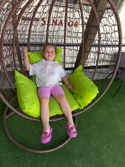 A young girl with blonde hair pulled back is seated in a hanging wicker chair with bright green cushions. She is wearing a light grey t-shirt with the words "JUST DO IT" in colorful lettering and pink biker shorts. Her feet are adorned with pink strawberry-themed sandals. The chair is suspended outdoors, with artificial green turf visible on the ground. In the background, a white caravan with red lettering reading "SENATOR" is partially visible. The scene appears to be set in a recreational area, possibly within a campsite or resort. The overall impression is one of a relaxed, sunny day.