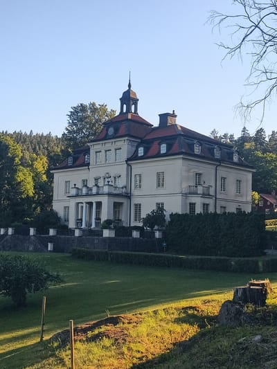 This media file captures a serene, sun-drenched scene at Skriketorp, Sweden. The focal point is a grand, two-story manor house, characterized by its pale yellow facade, red tiled roof, and a prominent central tower with a dark spire. The architecture features multiple windows with white frames, a covered balcony supported by columns, and dormer windows on the upper level.
Surrounding the manor is a meticulously maintained landscape. A low, dark stone wall with white decorative elements runs along the property line in front of the house, bordered by neatly trimmed green hedges. The foreground consists of a gently sloping, vibrant green lawn bathed in the warm, golden light of the late afternoon or early morning sun, casting long shadows. A weathered tree stump and a few rough rocks are visible on the right side of the frame.
Behind the manor, a dense forest of tall, green trees rises, creating a lush backdrop that contrasts with the clear, pale blue sky. To the far right, a small, red-tiled house with a chimney is partially visible, suggesting a residential area. The overall atmosphere is peaceful and tranquil, evoking a sense of calm and quietude. No people or specific activities are depicted. The time of day is clearly indicated by the low angle of the sun and the long shadows. The weather appears to be clear and pleasant.