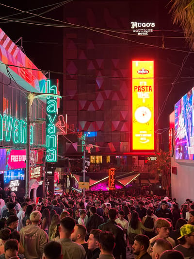 The image depicts a densely crowded street scene at night in what appears to be a nightlife district. The location is suggested to be Saint Julian's, Malta, based on the prevalence of businesses and signs such as "Havana," "Hugo's Hotel," "Clique," and "Jon Club," which are associated with the area.
A multitude of people are visible, predominantly facing forward, suggesting they are waiting or passing through the area. Many are looking down, potentially at their phones.
The street is flanked by buildings adorned with bright neon signs. "Havana" is prominently displayed in large green neon letters. A sign for "Hugo's Boutique Hotel" and an advertisement declaring "It's Pasta Time" are also visible. The signs are reflective, casting light onto the surrounding environment.
The background architecture has a geometric design and varying light fixtures. The overall atmosphere is vibrant and energetic due to the density of people and the bright lights. The presence of utility wires crisscrossing above the street is also evident.