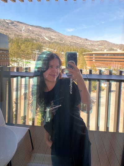 A woman is taking a selfie with a smartphone against the background of a mountain and a building in a city, possibly Озёрное, Russia. The view is seen through a glass window with multiple reflections. The woman is wearing a dark-colored shirt and a necklace. The city in the background contains buildings, trees, and a clear sky.