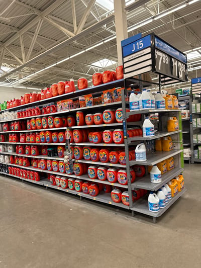 An aisle inside a large retail store, likely a supermarket or warehouse store in Worcester, United States, is filled with various laundry care products. The image, oriented vertically, shows multiple rows of shelves extending into the distance, packed with detergents and bleach.

The foreground features a blue aisle sign on a black frame, indicating "J15" and "Laundry Detergent." Below this, a price sign displays "$7.98" and "$13.98", with "Everyday Low Price" visible at the top.

The shelves are heavily stocked with distinct products. The upper shelves predominantly feature large yellow jugs of liquid detergent and white bottles of Clorox bleach with blue labels. Below these, the majority of the shelving space is dedicated to numerous orange containers of Tide Pods, stacked neatly in rows and columns. There are also larger orange jugs of liquid laundry detergent, possibly from the Tide brand, interspersed among the pods and on lower shelves.

The store's environment is characterized by a high, industrial-style ceiling with exposed metal trusses and bright fluorescent lighting fixtures. The floor is a plain, light-colored concrete. No people are visible in the aisle, suggesting a quiet moment or an uncrowded shopping area. The overall scene depicts a typical, well-organized retail display of household cleaning supplies.