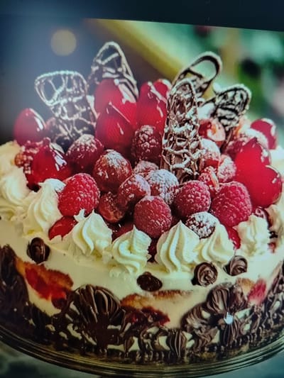 The image shows a close-up view of a richly decorated cake. The cake is covered in white whipped cream and adorned with fresh raspberries, red cherries, and elaborate chocolate decorations. There are also delicate chocolate curls and pieces dusted with powdered sugar. The sides of the cake are decorated with more intricate chocolate detailing that resembles foliage or waves. The background is softly blurred, with hints of green and yellow, suggesting an indoor setting perhaps a bakery or a dining area. There are no people visible in the image. The location of Yashkino, Russia is not depicted or suggested in the image.