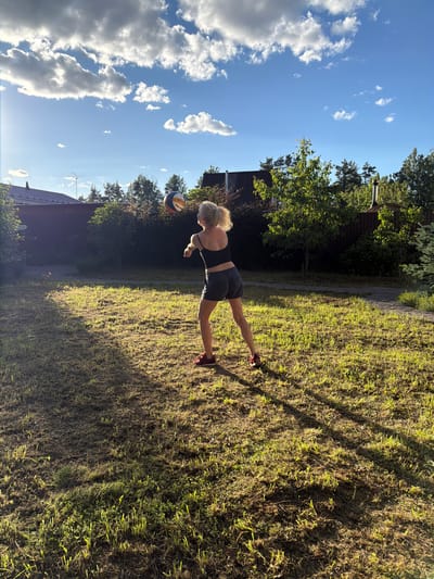 In Kerro, Russia, a woman with blonde hair tied in a ponytail is captured in mid-action, playing volleyball in a backyard. She is wearing a black sports bra and grey shorts, with red athletic shoes on her feet. Her body is angled towards the right of the frame, with her right arm extended upwards, about to hit a volleyball. The volleyball, with its distinct orange, blue, and white panels, is visible in the air above her.

The setting is a grassy yard, bathed in the warm, angled light of the late afternoon sun, casting long shadows across the lawn. The grass appears somewhat dry and uneven, with patches of new growth. Behind the woman, a dense hedge and trees form a natural backdrop, with the roof of a house visible to the left, partially obscured by foliage. The sky above is a vibrant blue, dotted with dramatic, fluffy white clouds, suggesting fair weather. The overall impression is one of casual outdoor recreation on a pleasant summer day.