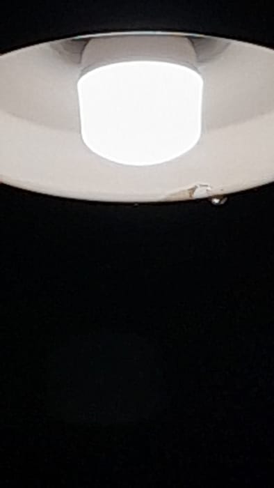The image shows a ceiling light fixture with a white, circular light bulb in a black room. The coordinates -34.667, -58.733 place the scene in the city of Merlo, Argentina. The image is a close-up of the light fixture, and there are no people or other objects in the image. The light bulb is turned on and illuminating the black room.