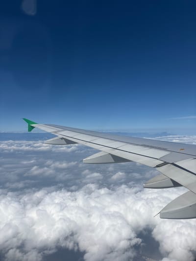 The image is an aerial view from an airplane, showing the wing of the aircraft extending into the frame. The wing is primarily white with a green tip. Below the wing, a vast expanse of white, puffy clouds fills the lower portion of the image. The sky above is a clear, deep blue, suggesting daytime. In the distance, a hazy, mountainous landscape is visible near the horizon line, dividing the sky and the clouds. The photo appears to be taken during daylight, possibly mid-morning or afternoon. The scene conveys a sense of travel and the vastness of the sky. No people are visible. There is no legible text in the image.