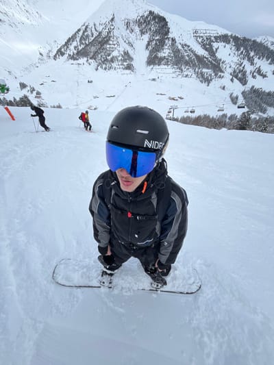 A person in snow gear, including a black helmet and blue goggles, is snowboarding on a snowy mountain. The person is facing the camera. In the background, snow-covered mountains are visible, and a ski lift runs along the mountain. Other skiers are visible in the background as well. The image appears to be taken at a high angle.