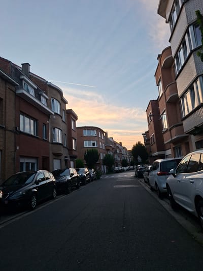 The image captures a street scene in Evere, Belgium, during a tranquil sunset. The sky is a soft canvas of pale blue with wispy clouds tinged with hues of orange and yellow, suggesting the golden hour. A faint contrail from an aircraft cuts a sharp line across the sky.
The street is lined with parked cars, predominantly dark-colored, casting long shadows on the asphalt. The vehicles appear to be modern sedans and hatchbacks. In the foreground, a few cars are more distinctly visible, with their reflections shimmering in the fading light.
On the left side of the street, a row of apartment buildings stands tall. Their facades are a mix of reddish-brown brickwork and lighter-colored detailing, featuring multiple windows that reflect the sky and surrounding structures. Some buildings have a curved architectural element, adding visual interest. The architecture suggests a residential urban environment.
The overall mood of the image is peaceful and serene, evoked by the soft lighting and quiet streetscape. The time of day is clearly late afternoon or early evening, marked by the sunset colors in the sky. No people are visible, contributing to the calm atmosphere. No text is discernible in the image.