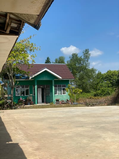 A bright, sunny day is depicted in Handil Baru, Indonesia. The image features a small, green building with a brown, tiled roof. The building has several windows with white frames and grates, and a double door entrance. In front of the building is a large, paved area. To the left, a portion of another building's roof and ceiling is visible, showing some signs of wear. The background consists of lush green trees and foliage under a clear blue sky with a few scattered white clouds. Some debris and plants are scattered around the base of the green building and the edge of the paved area. The scene appears to be an outdoor space, possibly a school or community building, in a tropical environment.