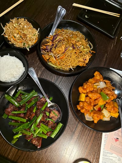 The image shows a table setting in a restaurant, featuring an assortment of Asian-style dishes. The dishes include chow mein, sweet and sour chicken, stir-fried beef with scallions, fried rice, and a bowl of white rice. Silver serving spoons are placed in each dish, except the bowls of fried and white rice. There are chopsticks on a small black square plate. A menu is partially visible in the upper right corner. The scene is located in a restaurant in Chula Vista, California.