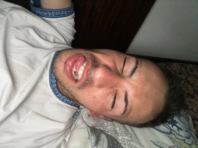 A young man with light skin and dark hair is lying on his side on a bed, looking towards the camera. He is wearing a white t-shirt with a blue and white patterned trim around the neck. His eyes are closed, and his tongue is sticking out. The background shows a dark wooden headboard on the left and a textured light-colored wall on the right. The bed has a patterned duvet. The image appears to be taken in a bedroom, likely in a home. The specific location within Popintsi, Bulgaria, cannot be determined from the visual information in the image.
