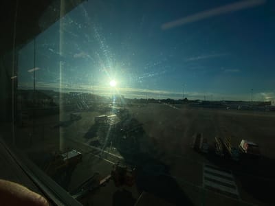 This image shows a view through a window, likely at an airport. The sun is setting or rising, casting bright rays and creating lens flare on the window, which also appears to have smudges or scratches. Outside, an airport tarmac is visible with several passenger boarding stairs, service vehicles, and distant aircraft. The sky is a clear blue with some wispy clouds. The scene appears to be from Luton Airport in the United Kingdom, given the provided context.