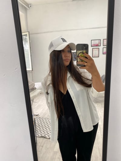 A young woman stands in front of a mirror, taking a selfie in what appears to be a room with white walls and light-colored wooden flooring. She is wearing a white mesh trucker hat with black lettering, an open white button-up shirt over a black V-neck top, and black pants. She has long, dark brown hair and is wearing a ring on her left hand. In the reflection, we can see a window on the left, and several framed certificates or pictures on the wall behind her. A small security camera is visible near the ceiling in the reflection. A patterned rug is on the floor. The scene is devoid of any specific Akyar, Russia, indicators.