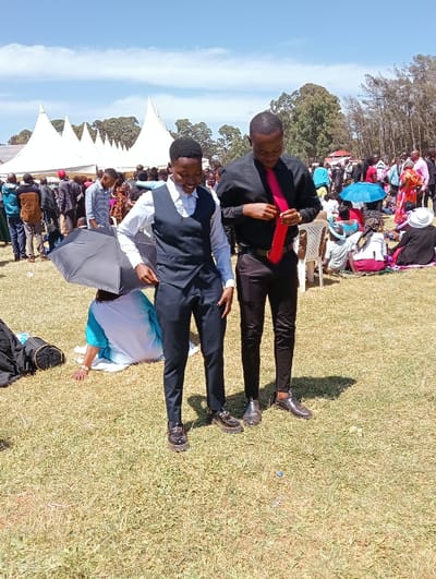 This image captures an outdoor gathering in Kimumu ward, Kenya, under a bright, sunny sky. The scene is set on a grassy field with numerous people attending what appears to be a community or social event.
In the foreground, two young men are the primary subjects. The man on the left wears a white long-sleeved shirt, a dark blue or black vest, and dark trousers, paired with dark formal shoes. He has a slight smile and appears to be adjusting his vest. Beside him, the man on the right is dressed in a black long-sleeved shirt and dark trousers, with a vibrant red tie that he is in the process of adjusting. He also wears dark formal shoes. Strong sunlight casts clear shadows behind them, indicating it is midday or early afternoon.
In the mid-ground and background, a large crowd of people is visible. Several white, pointed event tents are set up on the left side of the frame, suggesting a formal or semi-formal occasion. Many attendees are standing, while others are seated on the grass, some seeking shade under umbrellas, including a bright blue one on the right. A person in a light blue and white garment is seated on the ground behind the left man, with a black umbrella resting on the ground nearby. Trees line the horizon, completing the outdoor setting. The overall atmosphere suggests an active and well-attended event. No visible text is present in the image.