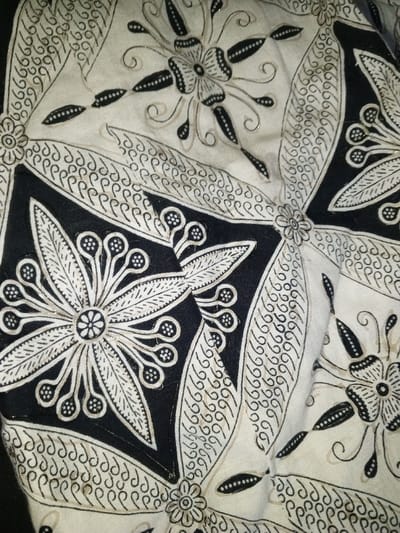 A close-up view depicts a textile with a black and off-white/cream batik-style pattern, located in Na Sang, Laos. The pattern features a repeating tessellation of stylized floral and foliate designs. Prominent motifs include large, multi-petaled flowers with a central circular element, surrounded by elongated leaf shapes and clusters of small, circular "berry" structures. These elements are rendered in off-white/cream against black backgrounds, and inversely, black against off-white/cream fields. Interspersed and filling background areas are dense, repetitive scroll-like or curvilinear patterns. The fabric exhibits visible creases and folds, indicating a draped or used state. The off-white areas show a slight yellowish tint, and minor dark speckling is present across the surface, suggesting wear or age. No individuals, actions, or additional environmental details are discernible.