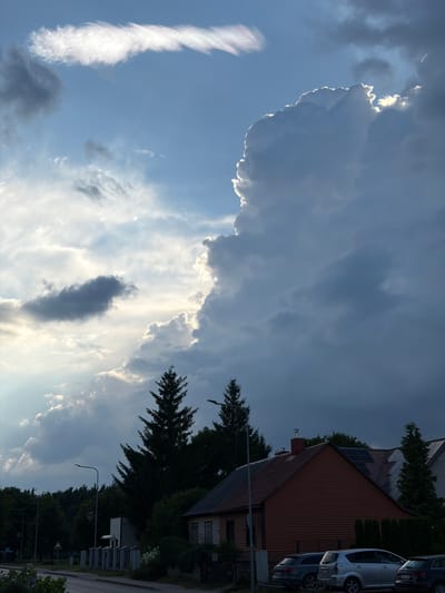 This is a landscape photograph taken in the city of Rūdiškės, Lithuania. The scene captures a dramatic sky filled with large, cumulonimbus clouds illuminated by the setting or rising sun. The sunlight creates a halo effect around the edges of the clouds, highlighting their contours. Below the sky, a residential area is visible, featuring houses with red-tiled roofs and solar panels. Several evergreen trees are scattered throughout the landscape, providing a contrast to the man-made structures. A street runs across the foreground, with parked cars along its side. The overall atmosphere is serene, with the natural beauty of the sky dominating the scene.