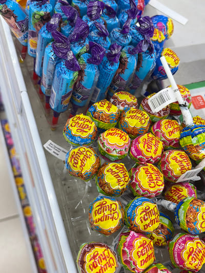 A close-up, slightly angled overhead shot shows a display of Chupa Chups lollipops in a retail setting. The lollipops are predominantly yellow with red and blue accents, featuring the "Chupa Chups" logo. Some lollipops are individually wrapped, with visible text indicating flavors like "strawberry" and "cola." A few lollipops are still in their original packaging, which is blue with cartoon characters and text. One of the lollipops has a barcode sticker attached. A white piece of plastic, possibly part of the display or packaging, extends from the upper right of the frame. A small white label with text in Indonesian, including an expiration date of "10/27 04/26", is visible on the lower left. The background is blurred, suggesting a store aisle. The overall impression is a well-stocked candy display. The image does not provide enough information to determine the specific location in Denasri Kulon, Indonesia, beyond it being a commercial establishment. There are no people or interactions visible in the image.