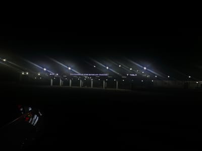 This media file captures a nighttime scene at an electric vehicle charging station in Dalvarzin, Uzbekistan. The foreground is dominated by the dark silhouette of a car, its surface reflecting the bright lights of the charging station. The car appears to have some visible branding or decals, though they are mostly obscured by darkness and reflection.

The main subject is the charging station itself, illuminated by rows of bright, white lights that create starburst effects and long streaks across the frame due to the long exposure. The structure of the station is visible, featuring a covered area with multiple columns and what appear to be charging bays behind a fence.

Text visible on the station reads "ELEKTROMOBILLAR UCHUN QUVVATLAH STANSIYASI," which translates from Uzbek to "Electric Car Charging Station." To the right of this main sign, the Tesla logo and the word "TESLA" are prominently displayed in illuminated lettering.

The time of day is clearly night, with the sky appearing completely black, indicating a lack of ambient light apart from the artificial sources. The weather appears clear, as there are no visible signs of rain or fog. The overall mood is stark and modern, with the bright, futuristic lighting of the Tesla charging station contrasting sharply with the deep darkness of the surrounding environment. The scene suggests a modern infrastructure development in an otherwise dark landscape.