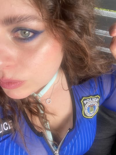 The image shows a close-up of a person's face and upper torso. The person has green eyes, brown hair, and is wearing blue eyeshadow. They are also wearing a light blue choker with a small circular pendant and a sheer blue top with a "POLICE" patch on the left chest. The background appears to be outdoors, with a hint of greenery and what looks like the slats of a bench or railing. The lighting suggests it is daytime. There is no information in the image that indicates the location in the city of Joensuu, Finland.