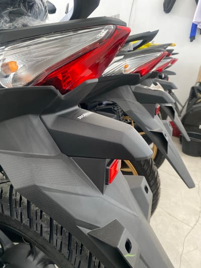 The image displays a close-up, low-angle view of the rear sections of several black motorcycles lined up in what appears to be a showroom or dealership. The motorcycles are facing away from the viewer, with their tail lights, fenders, and tires prominently featured. The tail lights are a combination of clear and red lenses, with integrated amber turn signals. The fenders are made of black plastic with a textured surface. The tires are visible, showing their tread pattern. A "HONDA" logo is visible on the fender of the foremost motorcycle. The background is blurred, showing white walls and possibly other motorcycles or merchandise. The lighting suggests an indoor setting. The scene is likely located within a commercial establishment in San Jose del Monte, Philippines, given the context of the image. There are no people or actions depicted.