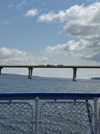 The image depicts a wide shot of a large bridge spanning across a body of water. The bridge has a flat, orange-colored deck with lights lining its edge, and it is supported by massive concrete piers. Several vehicles, including trucks, are visible on the bridge. The water is a deep blue, with small waves. In the foreground, a portion of a boat's railing is visible, made of grey metal mesh with a blue top rail. The sky above is light blue with scattered white clouds. The location is identified as the city of Oryol, Russia, specifically likely referring to the Oryol Bridge (also known as the Presidential Bridge) which crosses the Oka River.