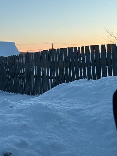This image depicts a winter outdoor scene, reportedly in the city of Bilimbay, Russia, at dawn or dusk. The foreground is dominated by undulating snowdrifts, appearing heavily textured. A dark, out-of-focus object is partially visible on the extreme right, suggesting the point of view. In the midground, a dark, weathered wooden picket fence, composed of vertical planks with visible gaps, extends across the frame. Snow is accumulated against the base of the fence and visible through some gaps. Beyond the fence, to the left, a portion of a building with a steep, snow-laden roof is visible. The background features a sky transitioning from a pale blue overhead to a soft orange-pink hue near the horizon. A utility pole with overhead power lines spans horizontally in the mid-distance. Sparse, bare tree branches are discernible on the far right against the sky. No human figures or active subjects are present.