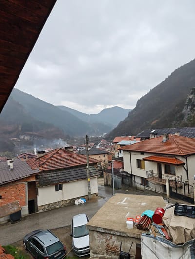 The image shows an elevated view of a portion of Grohotno, Bulgaria, on a cloudy, overcast day. A cluster of houses with terracotta-tiled roofs dominates the middle ground. In the foreground, parts of the rooftop of a building are seen, with construction supplies stacked on top: a red barrel, paint buckets, and metal tubes. Two cars are parked in the street below: a dark gray hatchback and a white sedan. In the background are rolling, forested hills, partially obscured by mist or low-hanging clouds. Some smoke is visible rising from the distant houses. Overhead power lines stretch across the scene. The composition appears to be taken from a building or higher vantage point.