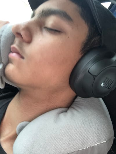 A young individual, likely male, is depicted sleeping in a close-up, angled shot. The subject's eyes are closed, and the head is tilted, resting on a light gray, U-shaped travel neck pillow. Dark hair is visible, along with a portion of a dark-colored baseball cap on the head. Black over-ear JBL headphones are worn, covering both ears. A dark-colored garment is visible beneath the neck pillow. The background is out of focus and indistinct, suggesting an indoor or vehicle environment. No visual information within the image identifies the location as Dettwiller, France, or any other specific geographical area.