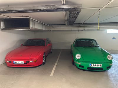 The image depicts a brightly lit underground parking garage in Espoo, Finland. Two classic Porsche sports cars are parked side-by-side. On the left is a vibrant red Porsche 944 with the license plate ZCU-757. To its right is a striking green Porsche 911 Carrera 2 (964 generation) with the Finnish license plate LZG-202. The cars are parked in designated spots marked by white lines on the concrete floor. Above the vehicles, the ceiling features extensive electrical conduits, ventilation systems, and lighting fixtures. A chain with yellow and black links hangs from the ceiling near the green Porsche. The overall environment is clean and well-maintained, characteristic of a modern parking facility.