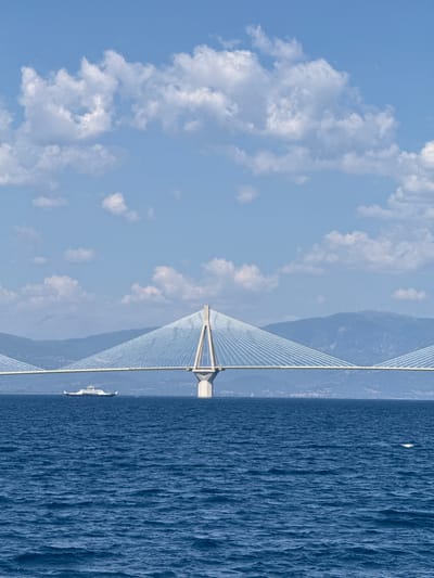 This is a vertical, eye-level, medium shot of the Rio-Antirrio Bridge in Patras, Greece. The bridge is a cable-stayed bridge that spans the Gulf of Corinth, connecting the Peloponnese peninsula with mainland Greece. The bridge is the main subject, dominating the center of the frame. It is a modern, impressive structure with a single concrete pylon supporting the deck, from which numerous white cables fan out to hold the bridge in place. The deck of the bridge is visible, stretching across the water, and a ferry boat is sailing beneath it. The water is a deep blue, with small whitecaps indicating a gentle breeze. In the background, hazy blue mountains rise, and the sky is a clear blue with scattered white clouds. There are no people visible in the image. The scene is peaceful and majestic.