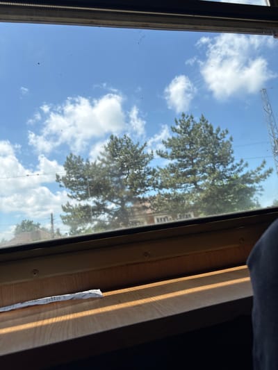 The image depicts a scene observed from inside a vehicle, likely a train or bus, looking out a window on a bright, sunny day. The sky is a clear blue with scattered white cumulus clouds. Two prominent, dark green pine trees are visible in the middle ground, with additional vegetation and residential structures, including houses with red-tiled roofs, discernible in the distance. On the far right, a partial view of a tall, metallic lattice structure, possibly an electricity pylon or telecommunication tower, is present. The window glass exhibits dust, smudges, and minor debris on its surface.

Internally, a light-colored wooden window sill or table is visible below the window pane. Parallel bands of sunlight and shadow are cast across this wooden surface, indicating direct sunlight entering the window. A small, crumpled piece of white paper or foil lies on the left side of the wooden surface. On the far right of the frame, a section of dark blue or grey fabric, consistent with human clothing (e.g., a leg or knee), suggests the presence of a passenger. The vehicle appears to be stationary or moving slowly through an environment consistent with the outskirts of a settlement. The described scene is situated in the city of Ivanovo, Bulgaria.
