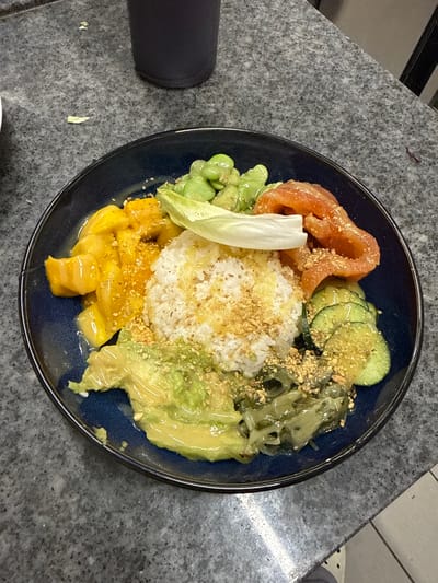 A close-up, overhead shot shows a dark blue bowl filled with a colorful poke bowl. The bowl is placed on a dark gray speckled countertop. The poke bowl is divided into sections. In the center, there is a mound of white rice topped with a light yellow dressing and sprinkled with chopped nuts. To the left of the rice are chunks of bright yellow mango. Above the rice and to the left are bright green broad beans. To the right of the rice are thin, curved slices of salmon. Below the rice are slices of cucumber and a green paste, likely avocado. A piece of white lettuce or endive is placed on top of the rice. The entire poke bowl is drizzled with a light yellow dressing and sprinkled with chopped nuts. The background shows the countertop extending towards a dark gray cup or container on the upper left and a black table leg on the right. The scene appears to be indoors, likely in a restaurant or home setting in Rivesaltes, France. No people are visible in the image.