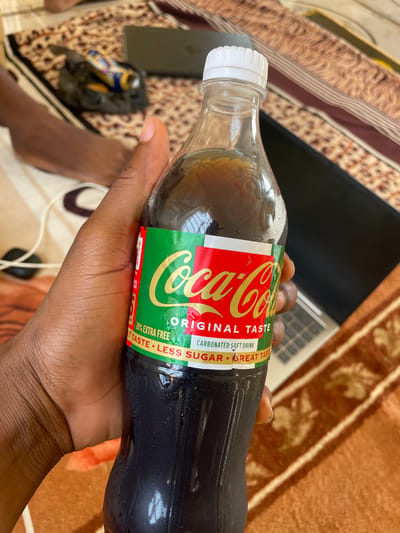 A hand holds a Coca-Cola bottle with a green and red label. The label reads "Coca-Cola," "ORIGINAL TASTE," and "20% EXTRA FREE CARBONATED SOFT DRINK," along with "GREAT TASTE • LESS SUGAR." The bottle is made of clear plastic and appears to be slightly condensation-covered. The background consists of a textured surface, possibly a rug or blanket, with a pattern of brown and beige. A laptop and a black object, possibly a phone, are visible in the upper right and left corners of the background, respectively. The lighting suggests it is daytime. The context provided states the location is Wai A, Nigeria.